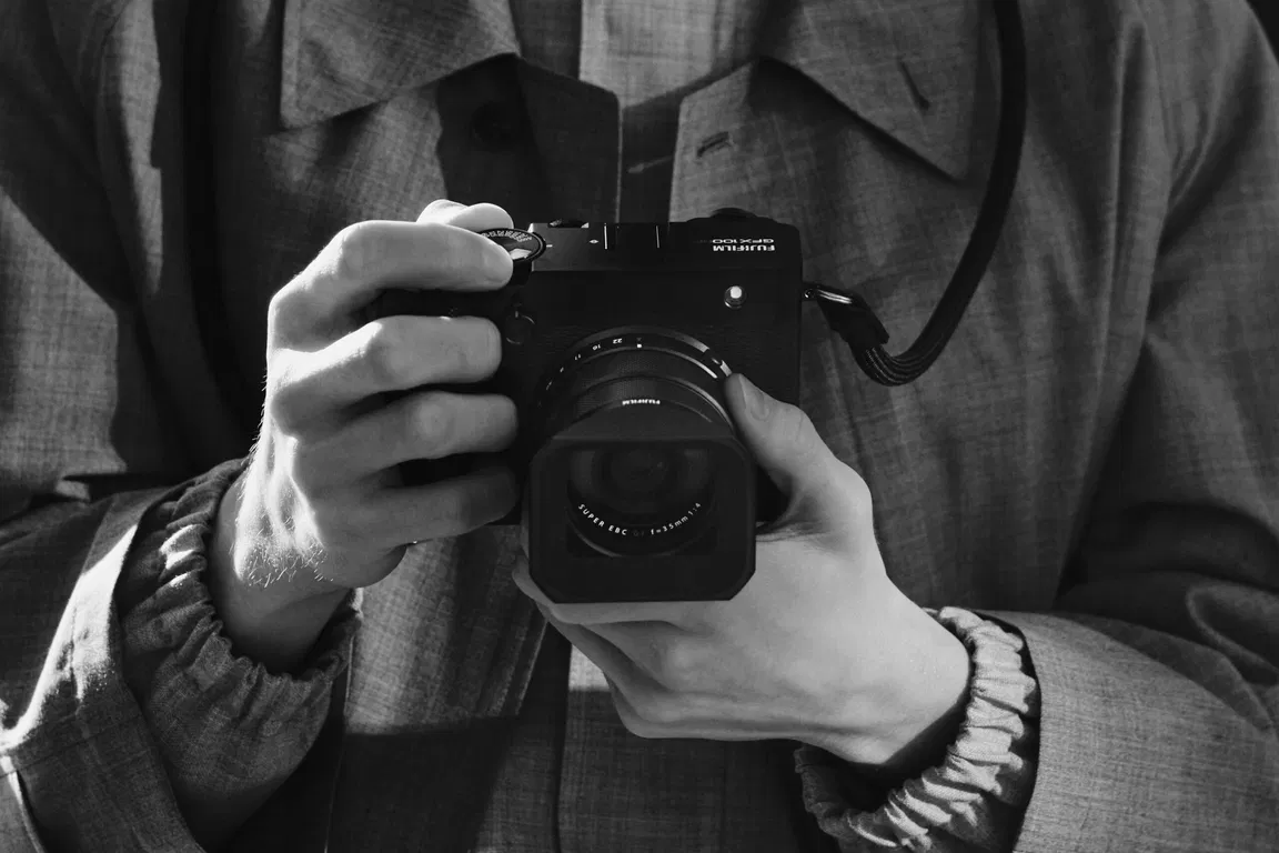 A person wearing a jacket holding a Fujifilm GFX100RF camera, focusing on the compact and lightweight design of the device.